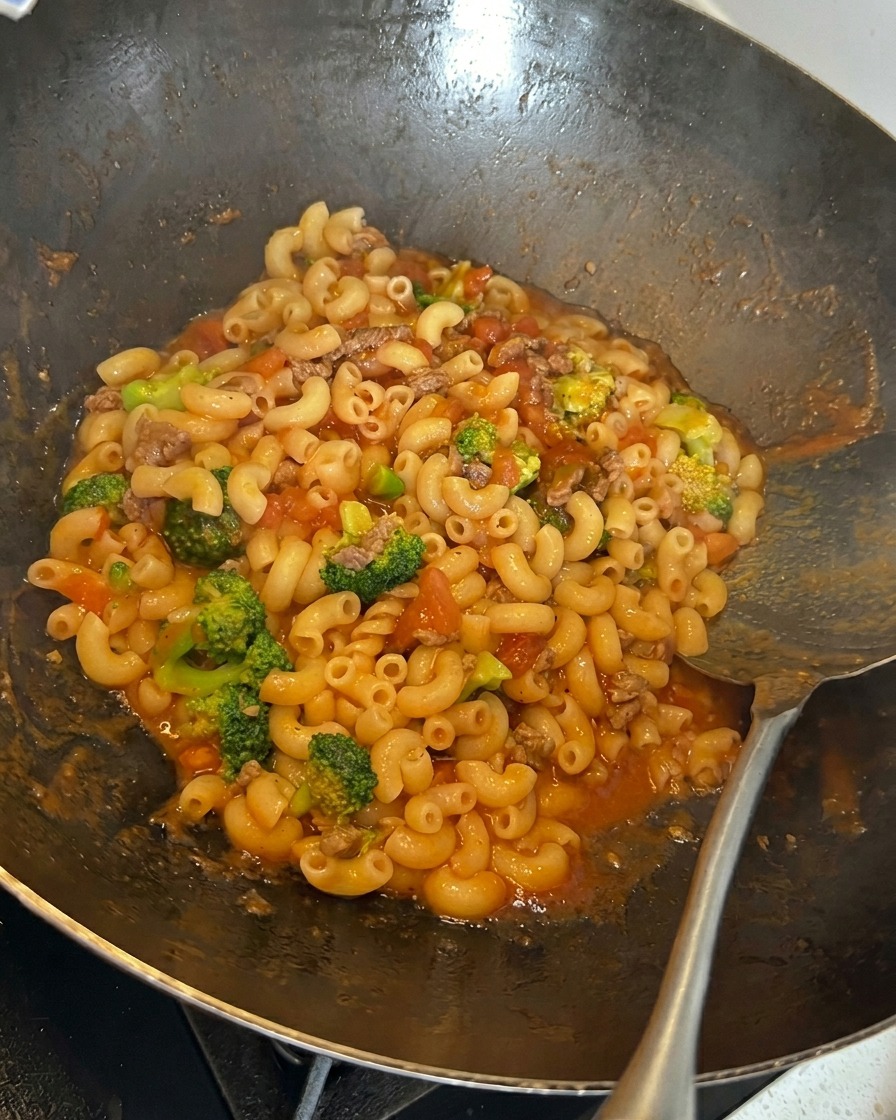 Queso rallado siendo espolvoreado sobre una mezcla de macarrones, brócoli y salsa de carne con tomate en un wok.