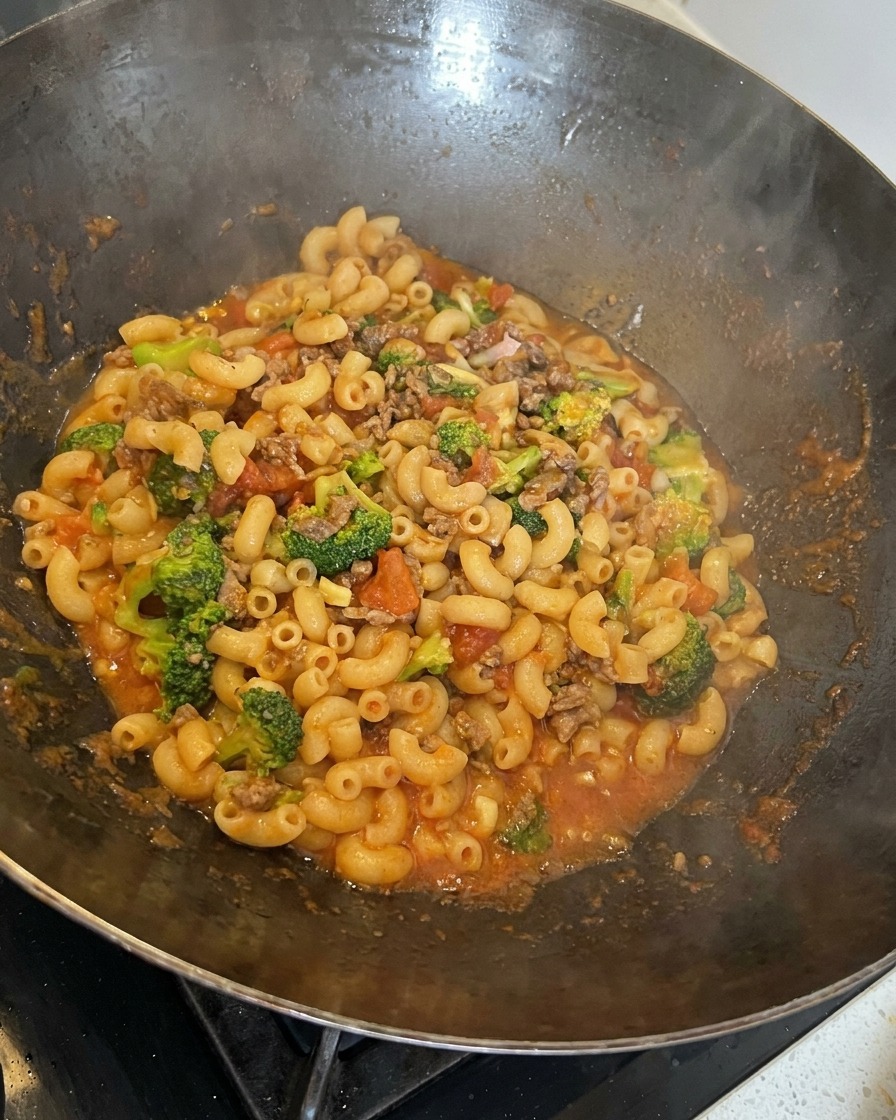 Pasta, ternera, brócoli y queso fundido siendo removidos juntos en un wok grande.