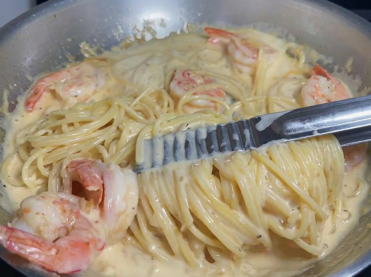 Pasta y camarones siendo mezclados con pinzas en una salsa cremosa para asegurar una cobertura uniforme.