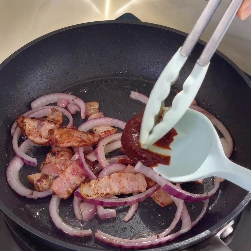 Salsa picante coreana siendo integrada con la mezcla de bacon y cebolla en la sartén.