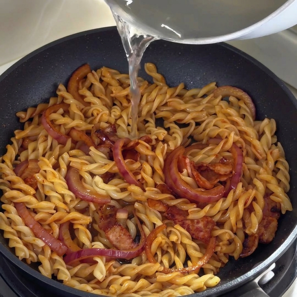 Agua de cocción caliente siendo vertida en la sartén con la pasta, el bacon y la cebolla.