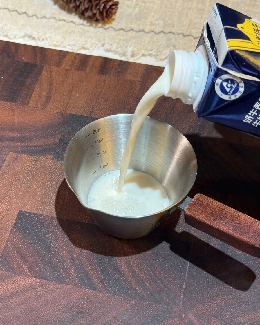 Leche blanca vertiéndose desde un brik azul en una taza medidora pequeña de acero inoxidable con mango de madera.