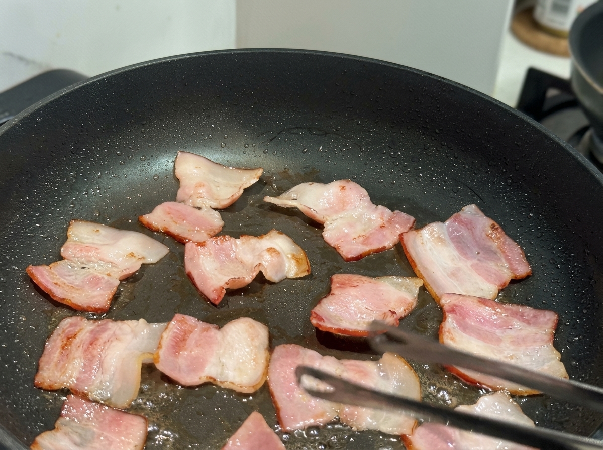 Pequeños trozos planos de bacon friéndose en una sartén antiadherente negra con unas pinzas visibles.