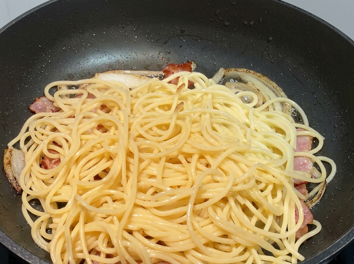 Espaguetis cocidos apilados sobre cebollas sofritas y trozos de bacon en una sartén negra.