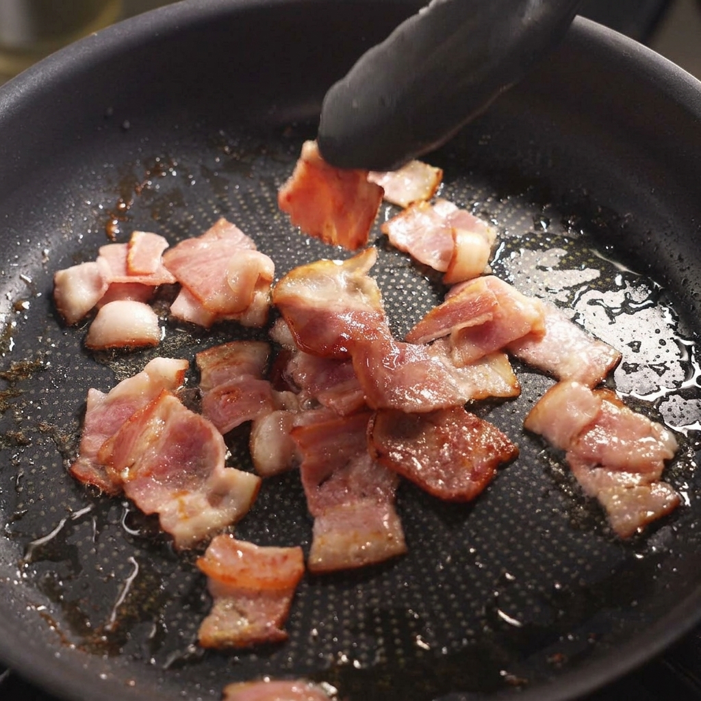 Unas pinzas negras dando la vuelta a unos trozos de bacon chisporroteantes y ligeramente dorados en una sartén.