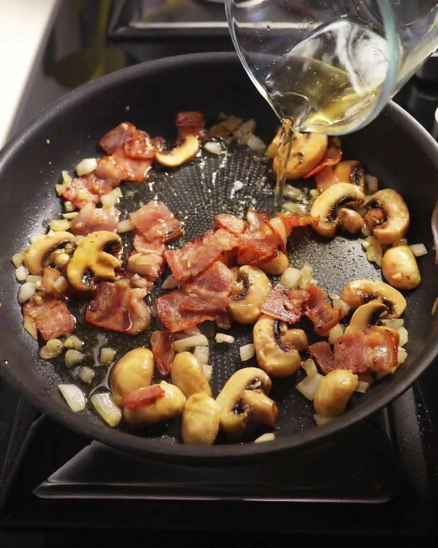 Vino blanco claro vertiéndose desde una jarra medidora de cristal en una sartén con champiñones y bacon cocinados.