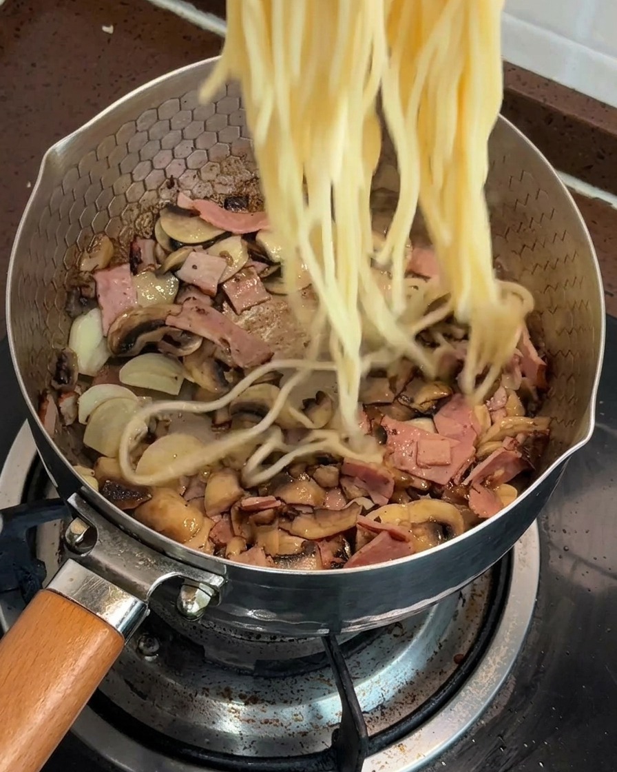 Espaguetis cocidos siendo añadidos a una sartén llena de una mezcla de champiñones y beicon salteados.