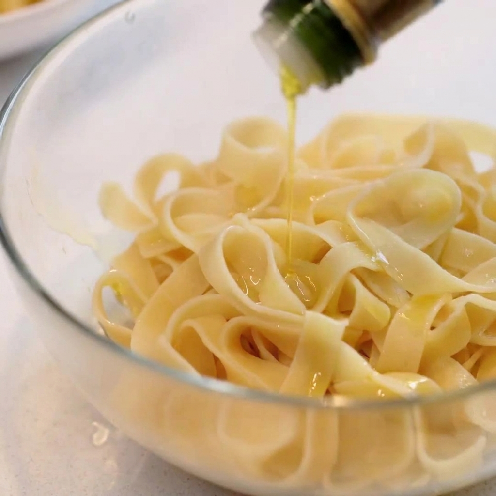 Pasta larga cocinada en un bol de cristal siendo rociada con aceite de oliva.