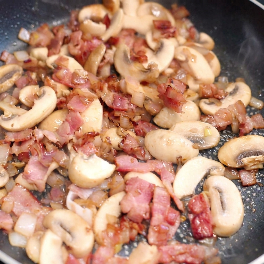 Pasta larga siendo mezclada en una sartén con bacon, champiñones y cebolla.