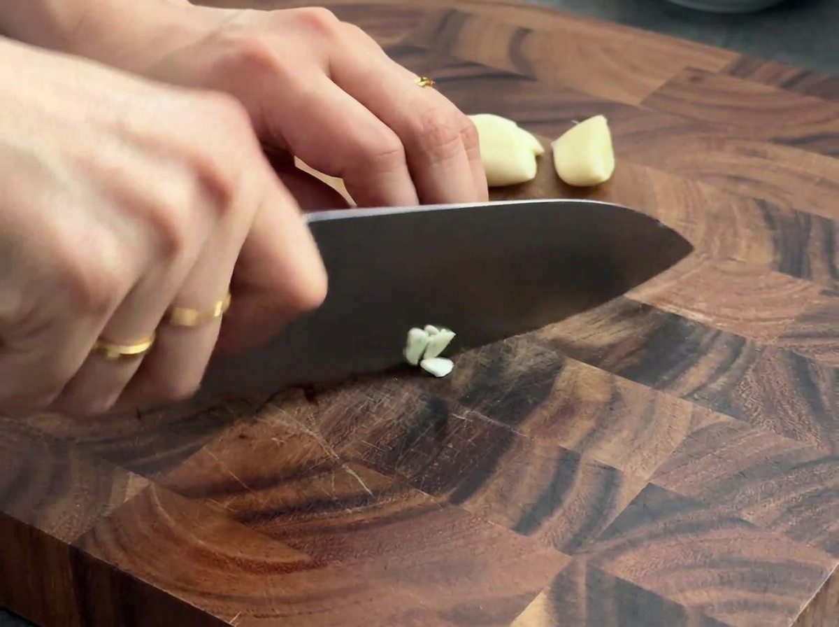 Dientes de ajo fresco siendo picados en una tabla de cortar de madera con un cuchillo de chef.