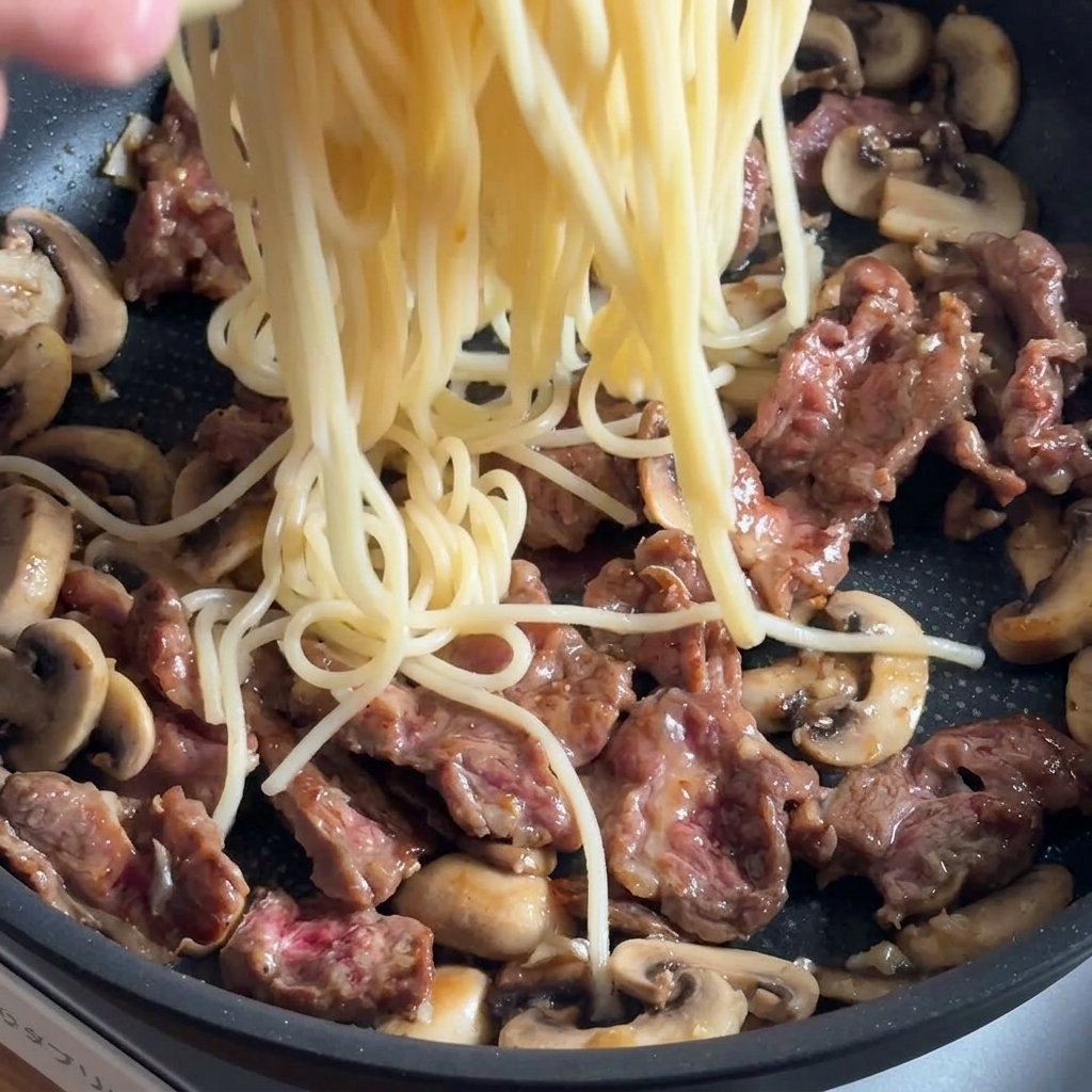 Pasta siendo añadida a una sartén con ternera y setas, lista para condimentar.