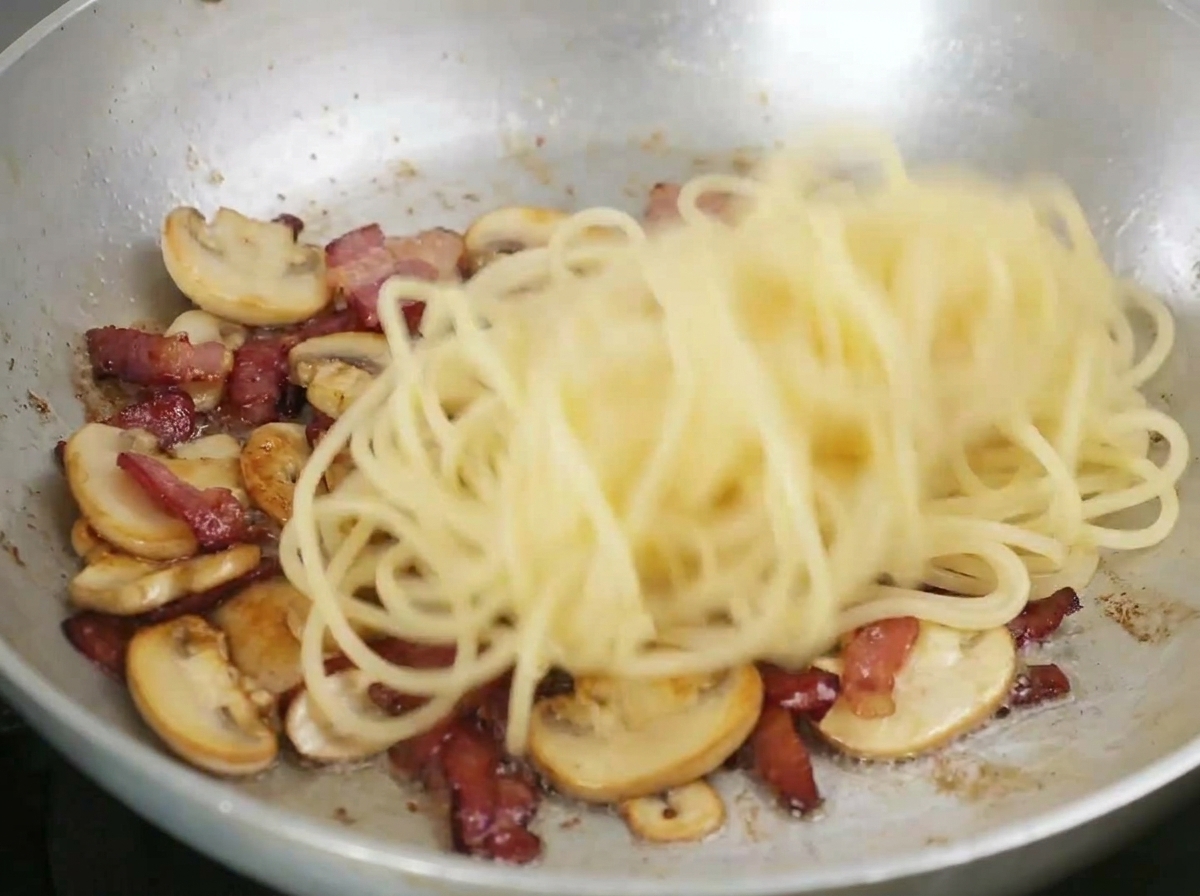 Espaguetis siendo salteados en una sartén con tocino frito y champiñones en rodajas.