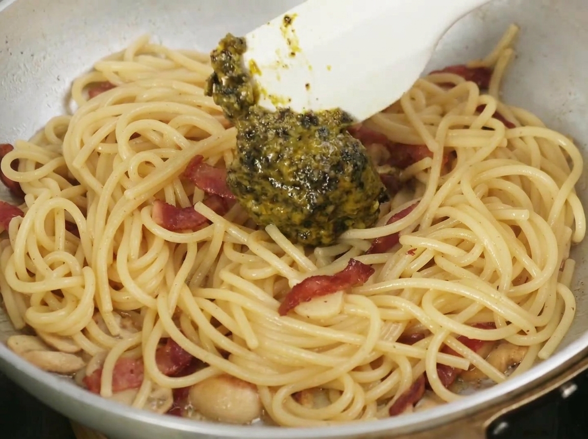 Una espátula mezclando la aromática y oscura salsa de trufa negra en una sartén con pasta cocida, ajo y tocino.