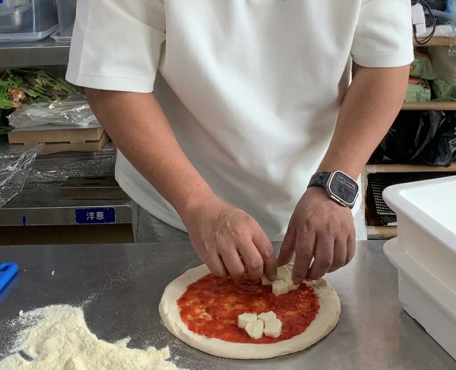 Manos colocando cubos de mozzarella fresca sobre una masa de pizza cubierta de salsa de tomate.