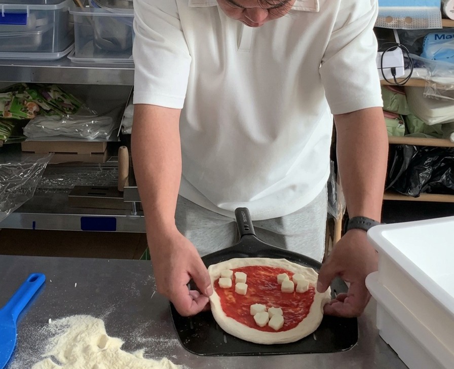 Manos ajustando los bordes redondos de una pizza Margarita sin hornear que descansa sobre una pala metálica perforada para pizza.