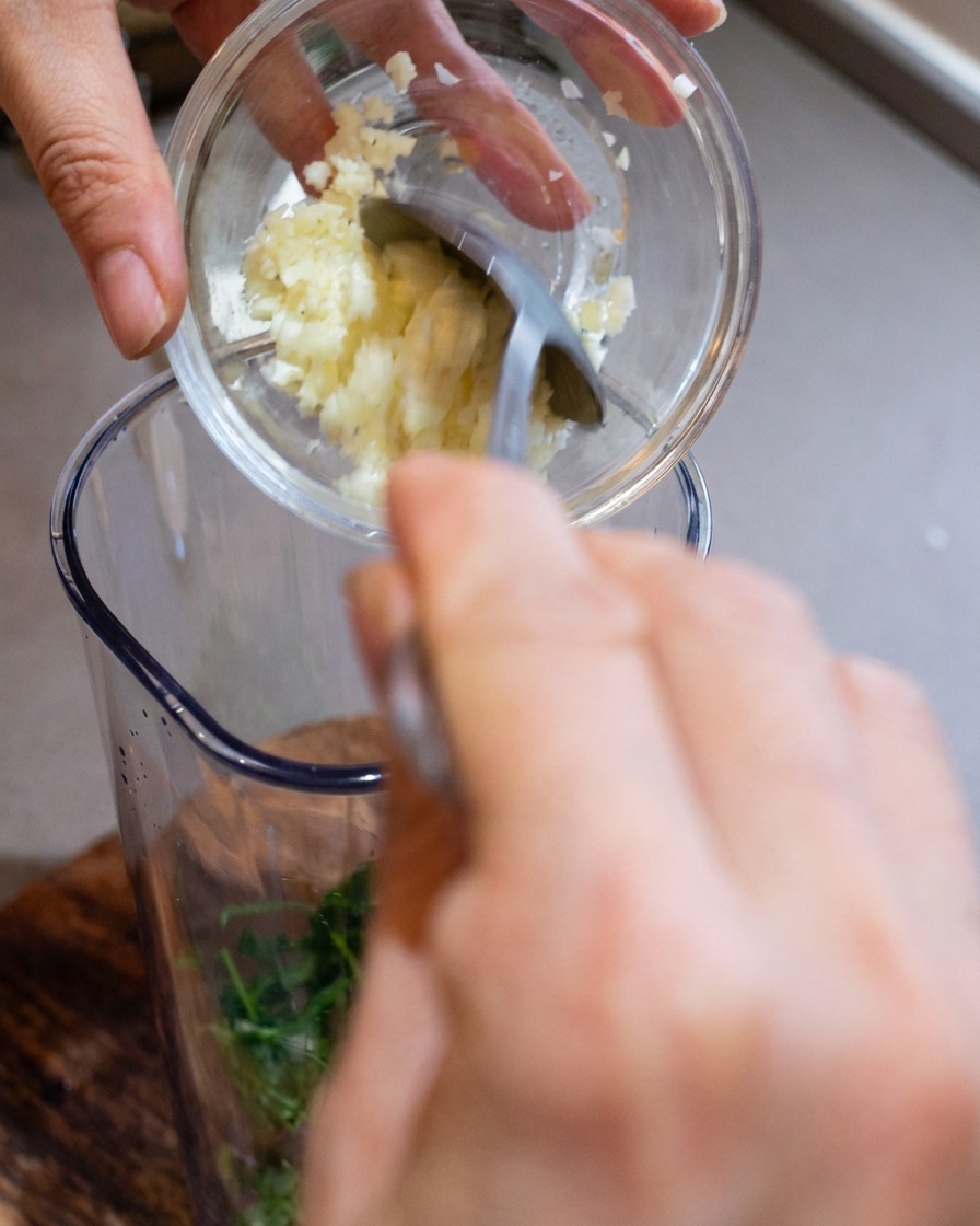 Un recipiente de licuadora siendo llenado con ajo picado, queso recién rallado y piñones tostados.
