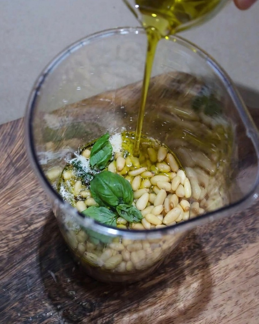 Una vista de cerca de ingredientes de pesto incluyendo albahaca fresca, piñones y parmesano siendo licuados en un recipiente con un chorrito de aceite de oliva vertiéndose.
