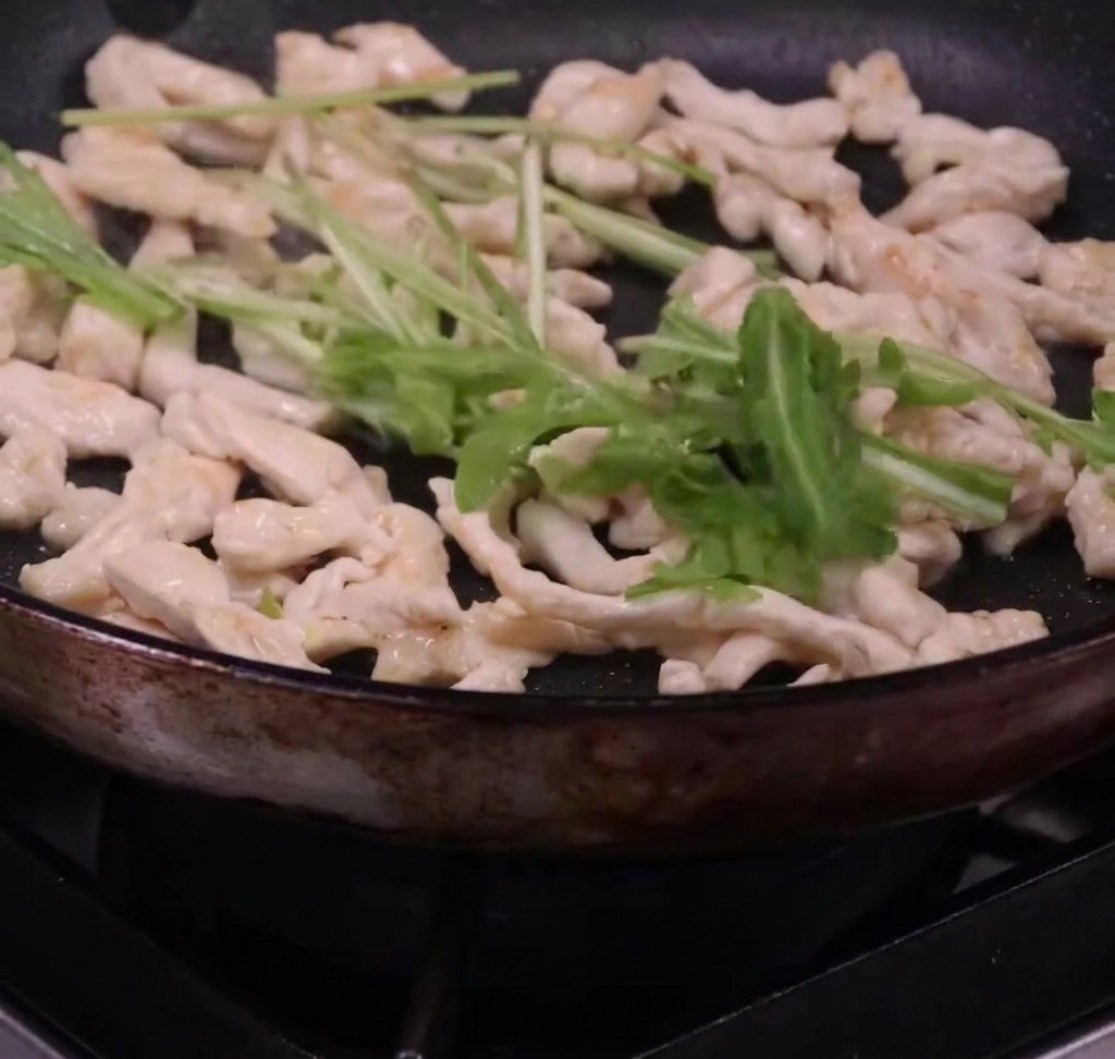 Espaguetis y rúcula fresca siendo mezclados con tiras de pollo cocinado en una sartén.