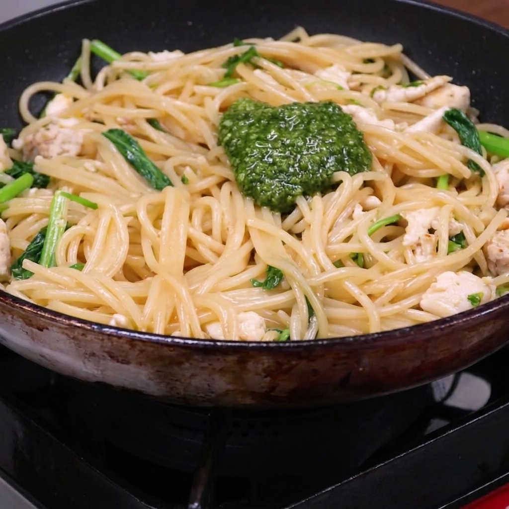 Una cucharada de pesto verde vibrante sobre una mezcla de espaguetis cocidos, trozos de pollo y rúcula en una sartén.
