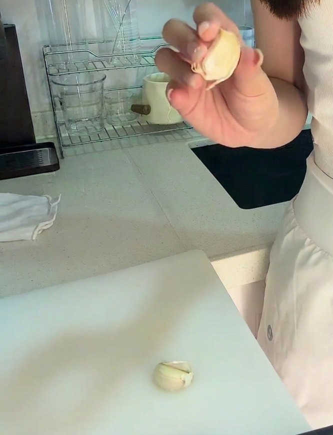 Una mano sosteniendo un diente de ajo pelado sobre una tabla de cortar blanca junto a un poco de ajo picado.