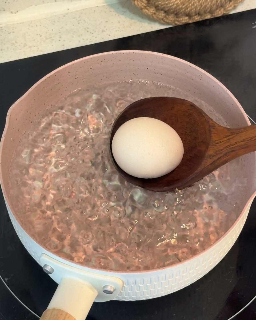 Un cazo de madera introduciendo suavemente un huevo entero en una olla rosa con agua hirviendo.