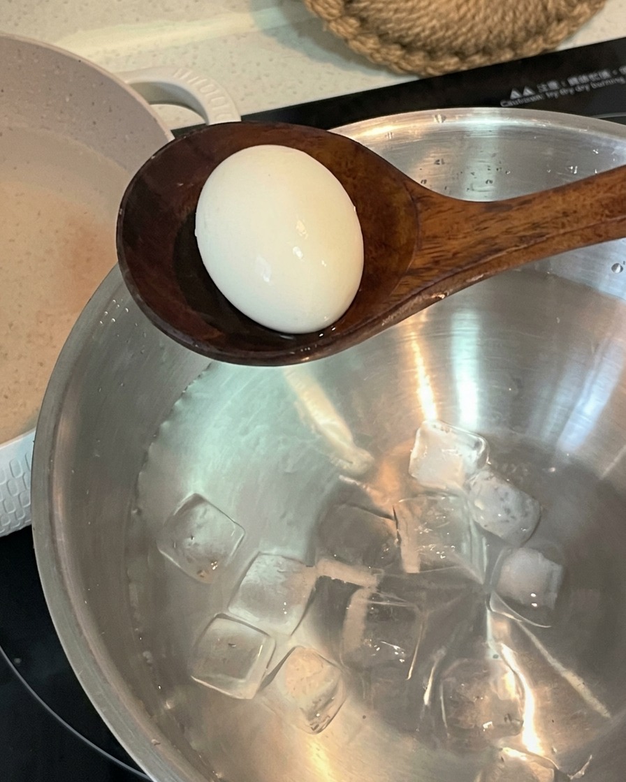 Una cuchara de madera transfiriendo un huevo hervido a un bol de acero inoxidable lleno de agua fría y cubitos de hielo.