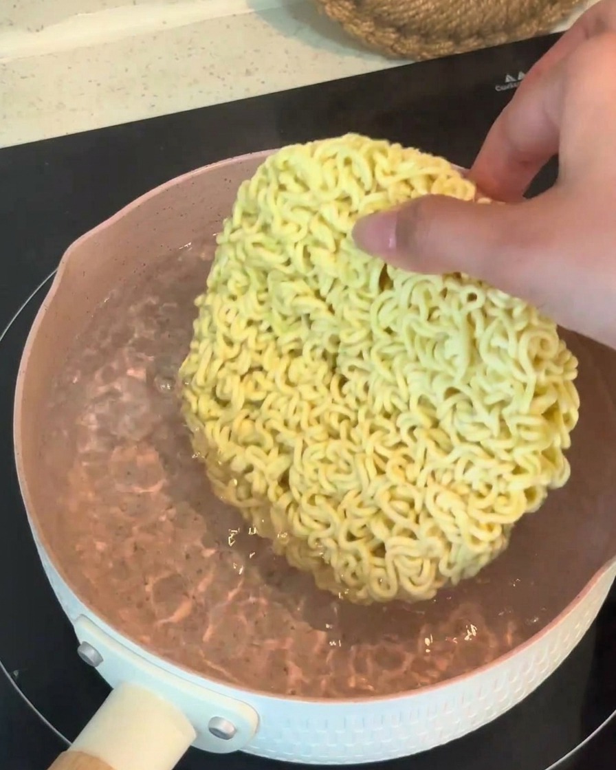 Una mano colocando un bloque seco y redondo de fideos ramen instantáneos en una olla de agua hirviendo.