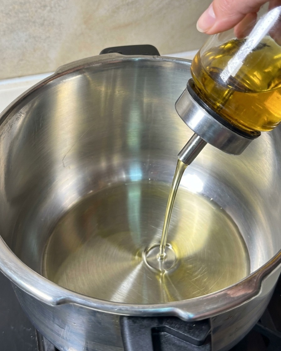 Aceite de cocina vertido desde un dosificador de cristal en una olla de acero inoxidable limpia.