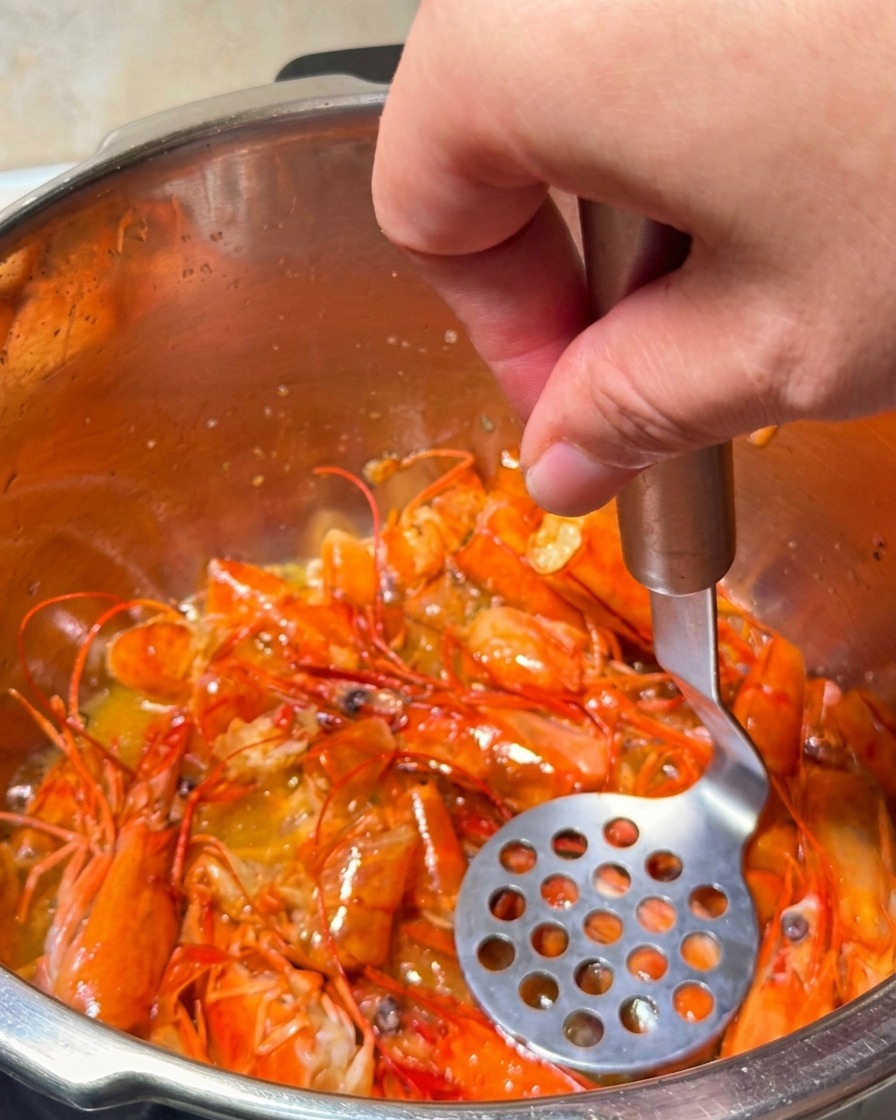 Un machacador de patatas presionando cabezas de gamba mientras se fríen para extraer aceite naranja dorado.