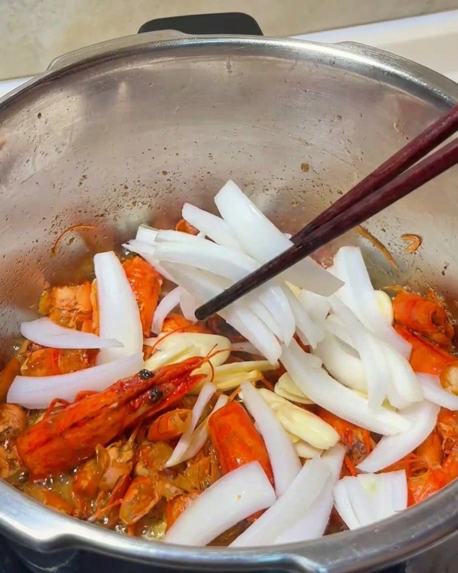 Primer plano de láminas de ajo y tiras de cebolla siendo integradas con las cáscaras de gamba fritas en una olla de metal.