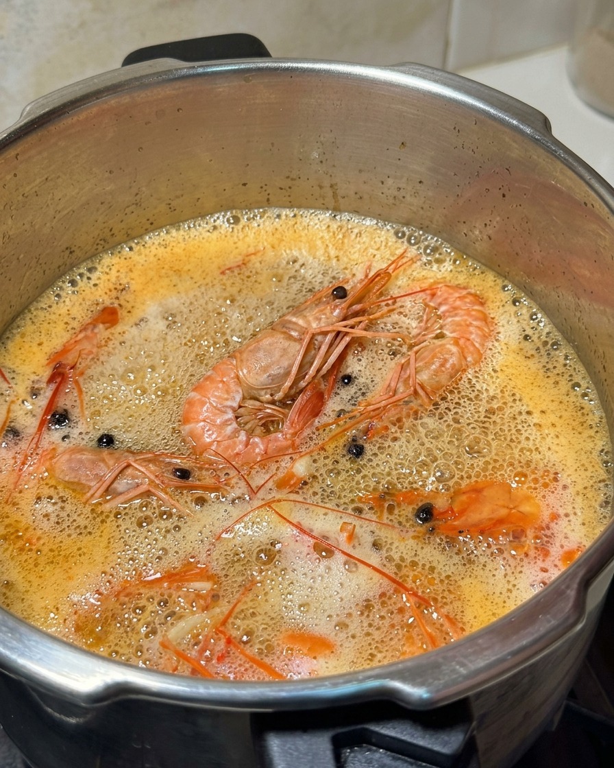 Un cucharón de metal retirando espuma blanca de la superficie de un caldo de gambas naranja hirviendo.