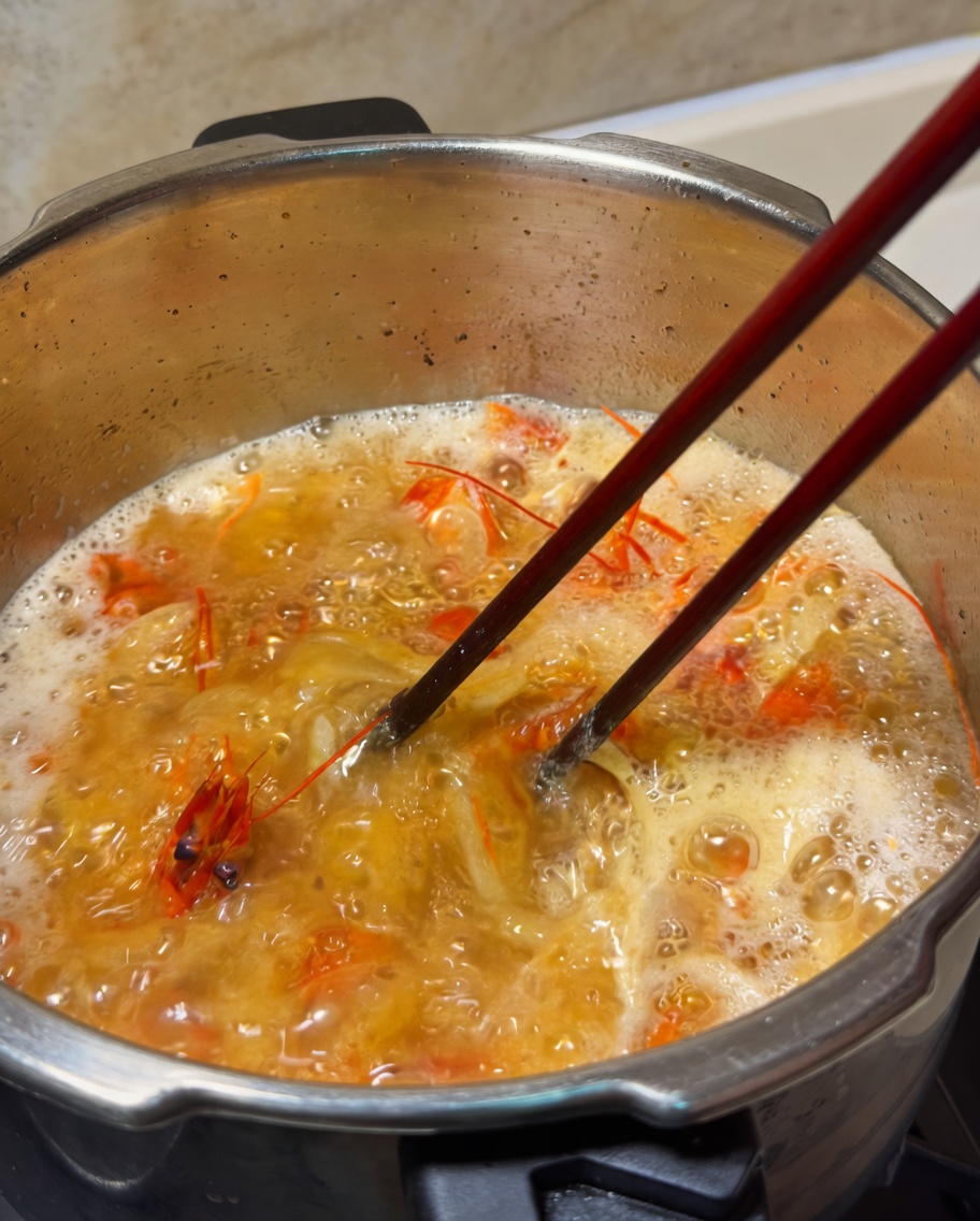 Una nuez de mantequilla derritiéndose en una olla de caldo de gambas naranja hirviendo, siendo removida con palillos.