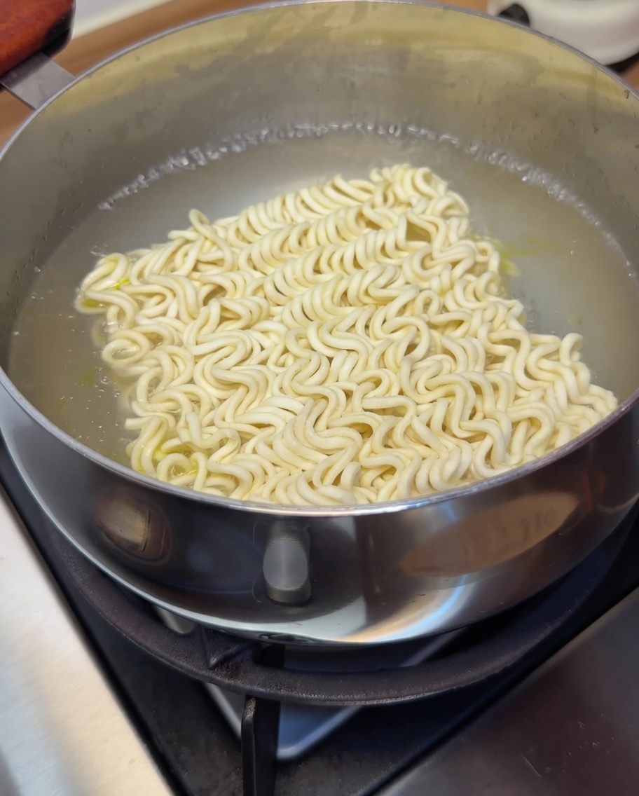 Un bloque de fideos instantáneos sumergidos en agua hirviendo, cocinándose hasta quedar tiernos.