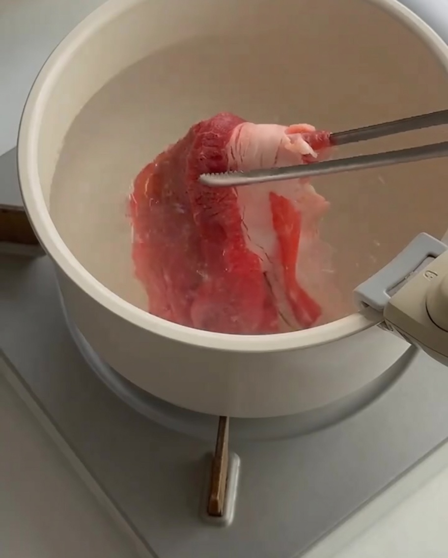 Finas lonchas de ternera cruda siendo sumergidas en una olla con agua hirviendo usando pinzas.
