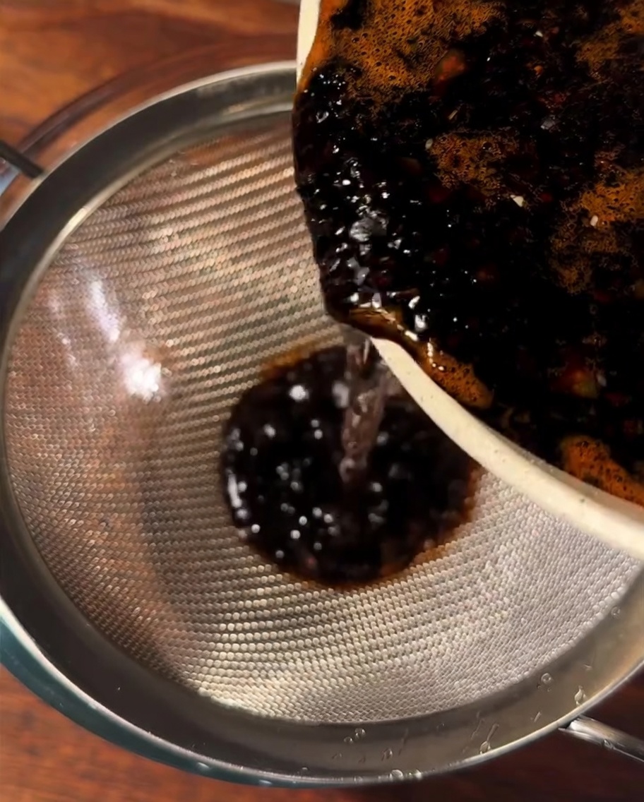 Salsa marrón oscura siendo vertida a través de un colador metálico en un bol de cristal.