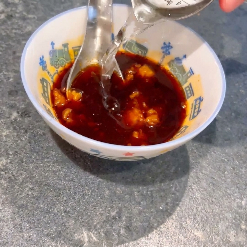 Vertiendo agua en un tazón pequeño que contiene salsa de soja oscura y grumos de miso, con una cuchara revolviendo la mezcla.