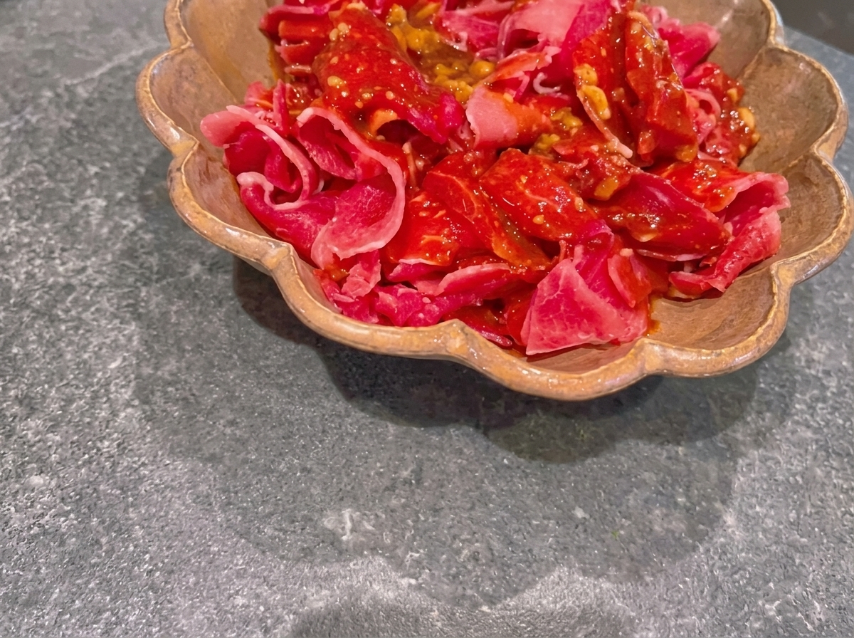 Finas rebanadas de carne de res cruda cubiertas con una marinada oscura de miso dentro de un plato marrón con bordes ondulados.