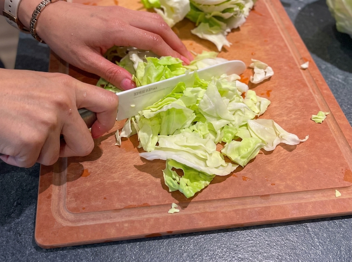 Manos usando un cuchillo de cerámica blanco para picar repollo verde fresco en trozos pequeños sobre una tabla de cortar de madera.