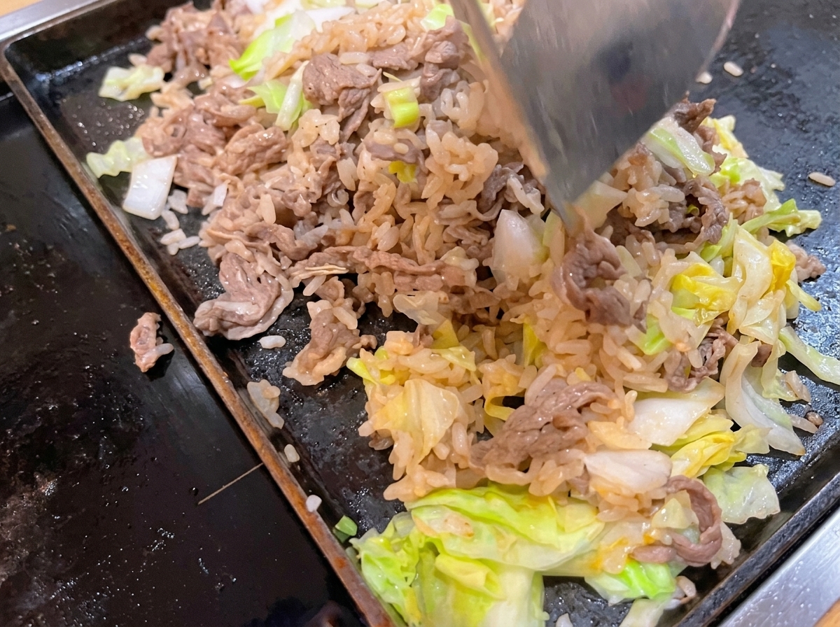 Una espátula de metal mezclando y revolviendo carne cocida, arroz blanco y repollo verde en una sartén plana caliente.