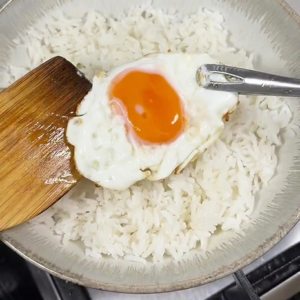 Espátula de madera colocando un huevo frito con la yema líquida sobre un bol lleno de arroz blanco.