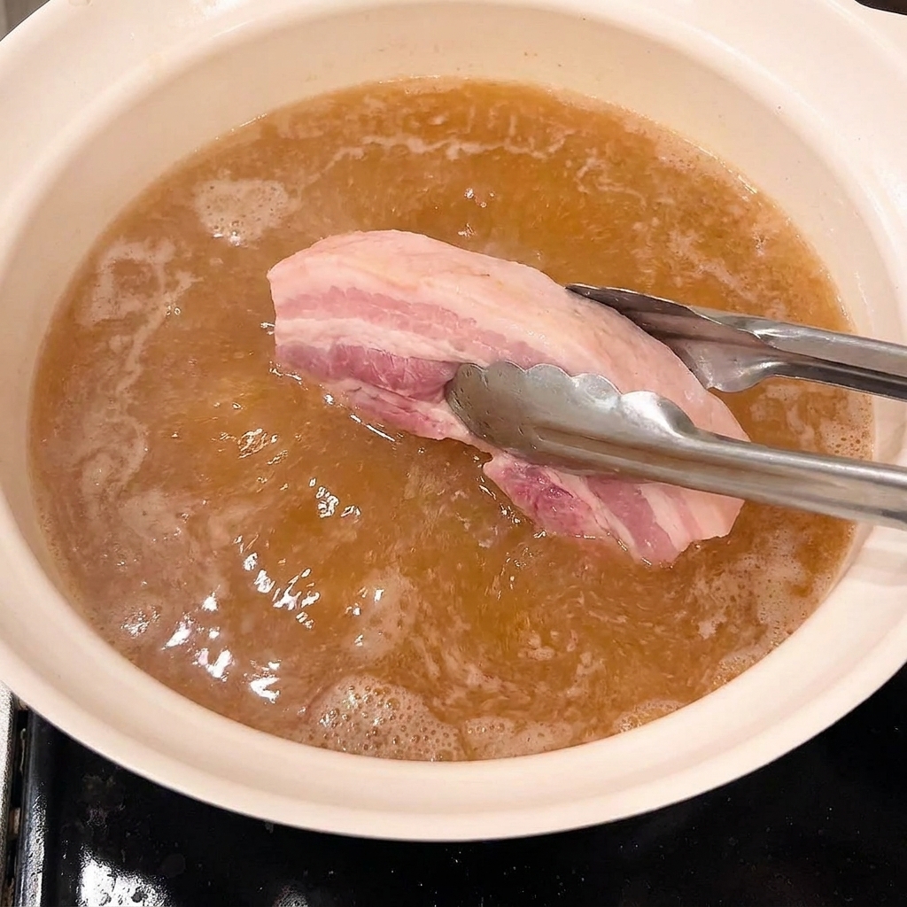 Pinzas colocando trozos de panceta escaldada en una olla con salsa de braseado hirviendo a fuego lento.