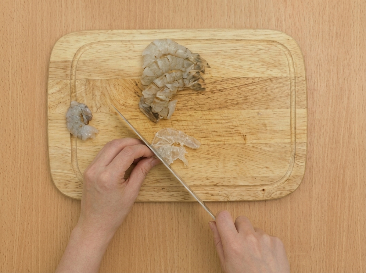 Primer plano de un cuchillo haciendo cortes superficiales a lo largo del vientre de un camarón crudo sobre una tabla de cortar de madera.