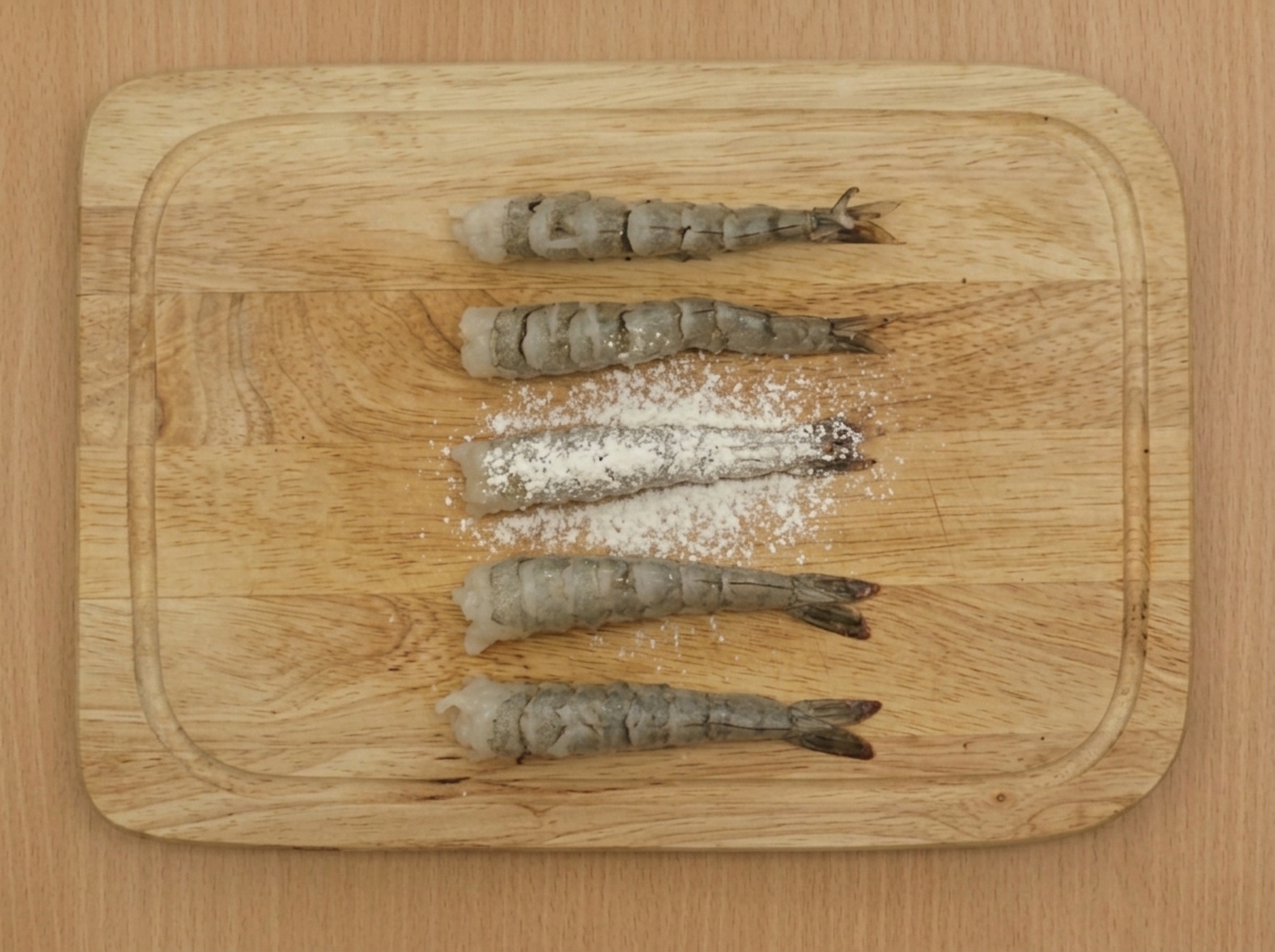 Un tamiz espolvoreando harina de bajo contenido en gluten sobre varios camarones preparados dispuestos en una tabla de madera.