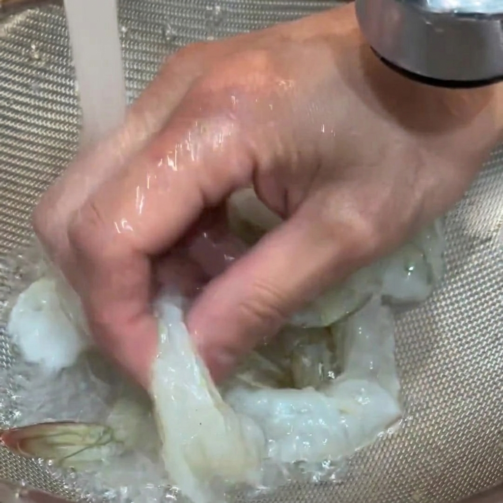 Camarones crudos siendo enjuagados bajo el grifo de agua fría en un colador.