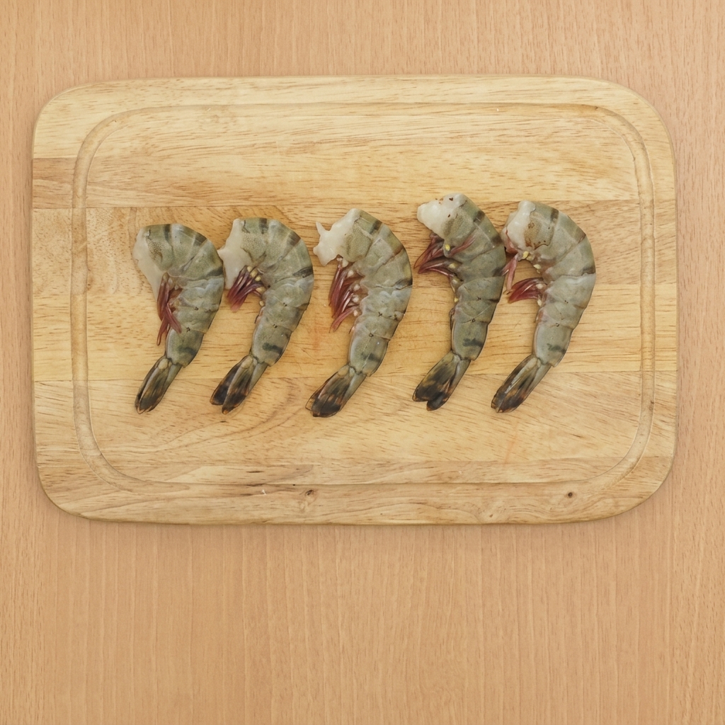 Cinco camarones limpios en una tabla de cortar de madera con las colas aún puestas.