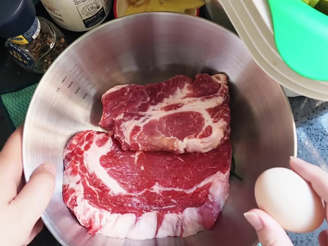 Un huevo crudo siendo roto sobre chuletas de cerdo frescas en un tazón de acero inoxidable.