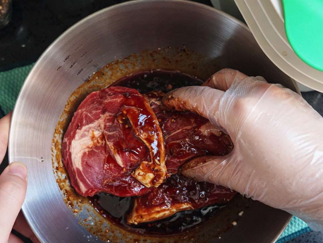 Mano con guante de plástico mezclando chuletas de cerdo crudas con el marinado en un tazón de metal.
