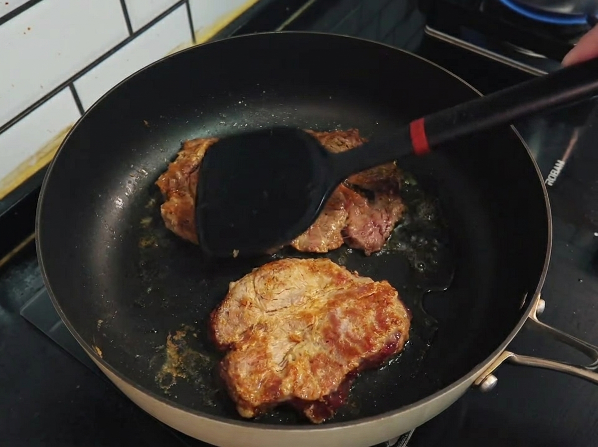 Espátula volteando chuletas de cerdo doradas en una sartén antiadherente.