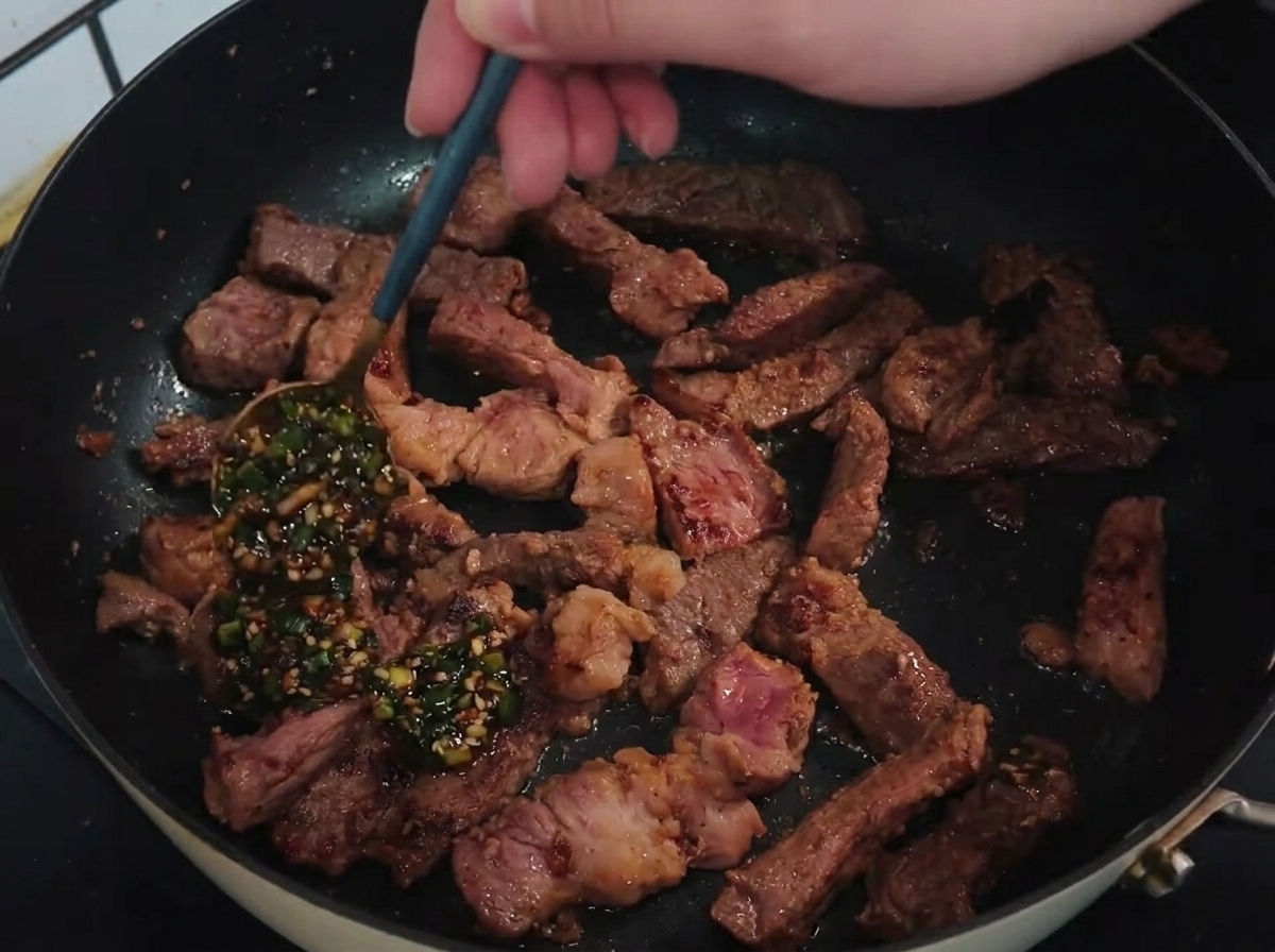 Cuchara vertiendo una salsa verde de cebollino y ajo sobre tiras de chuleta de cerdo selladas en una sartén caliente.