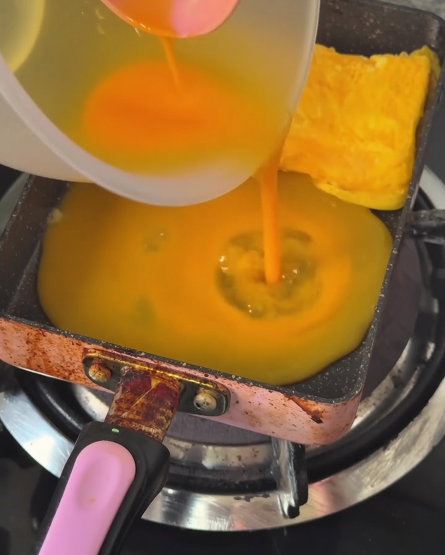 Una capa fina de huevo batido siendo vertida en una sartén rectangular caliente y engrasada.